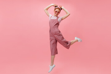 Lady in great mood jumping with raised hands on pink background. Portrait of girl with buns dressed in overalls and white T-shirt