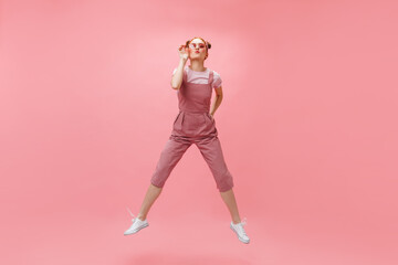 Fototapeta na wymiar Cheerful woman in overalls and white T-shirt puts on sunglasses and jumps on pink background