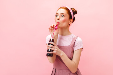 Lovely woman in bright summer sundress smiles and holds bottle with sweet soda on pink background