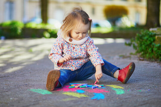Adorable toddler girl drawing with colorful chalks on asphalt