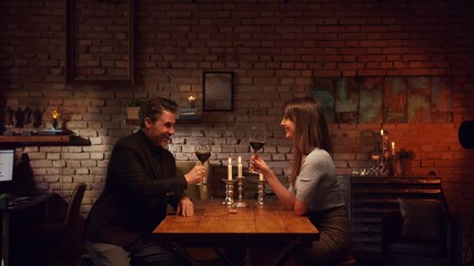 Elegant couple having romantic dinner at home, sitting at table in living room drinking red wine, talking. Stay at home concept.