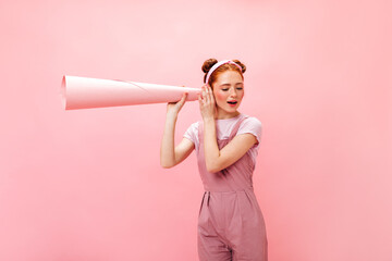 Young mischievous red-haired girl in pink jumpsuit eavesdrops using pink tube