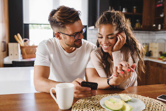 Beautiful Interracial Love Couple Using Smart Phone Having Good Time At Home In The Morning After The Breakfast