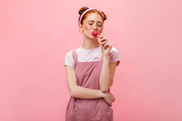 Young girl with buns dressed in pink dress eats lollipop on isolated background