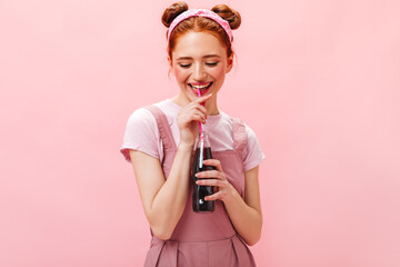 Portrait of cheerful red-haired girl sincere laughing on pink background. Woman in pink dress drinks sweet soda