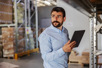 Young serious attractive bearded supervisor standing in warehouse and using tablet.