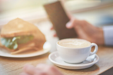 Energy drink. Cropped shot of a man using smartphone while having breakfast in cafe. Cup of coffee and fresh sandwich on wooden table