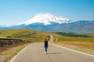 A woman walks along the highway against the backdrop of mountains. Freedom and travel concept. Elbrus, Caucasus.