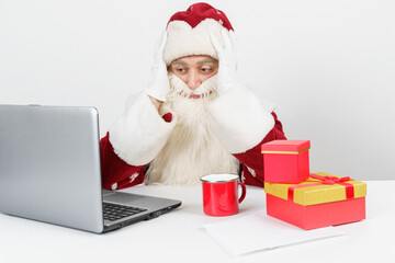 Tired Santa Claus sits at his desk, drinking coffee and working at the computer. There are Christmas gifts on the table.