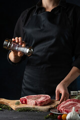 Close-up view of professional chef salts fresh beef or pork steak on wooden chopping board on black background. Backstage of cooking grilled restaurant meat. Food concept. Delicious meal for dinner.