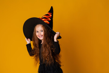 little girl with long red hair smiles in a Halloween witch costume and holds a pumpkin-shaped candy bowl on a yellow background. Halloween for children, space for text