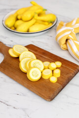 Sliced Yellow Crookneck Squash on a Wood cutting Board on Marble Countertop with a White Bowl of Uncut Squash in Background