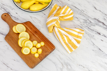 Sliced Yellow Crookneck Squash on a Wood cutting Board on Marble Countertop with a White Bowl of Uncut Squash in Background