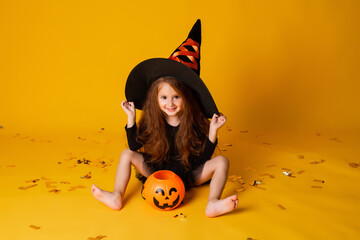 little girl with long red hair smiles in a Halloween witch costume and holds a pumpkin-shaped candy...