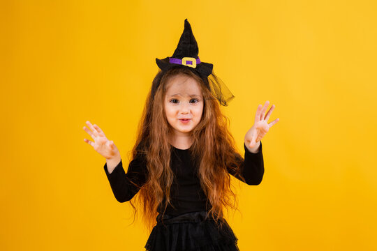 little girl with long red hair smiles in a Halloween witch costume and holds a pumpkin-shaped candy bowl on a yellow background. Halloween for children, space for text