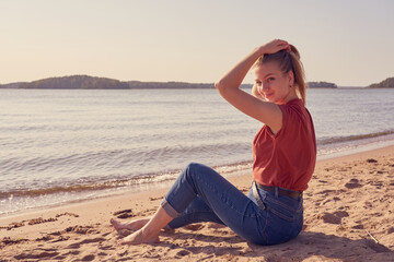 Fototapeta na wymiar Happy and beautiful young white woman is sitting on the sandy beach in sunlight in blue jeans and red shirt and lookin back. Copy space.