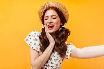 Adorable woman in boater and with bows on her hair shows her tongue and takes selfie on orange background