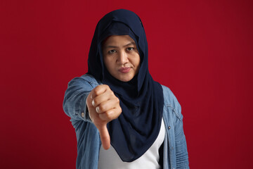 Portrait of muslim lady wearing hijab shows mocking face and thumbs down gesture, disappointed expression