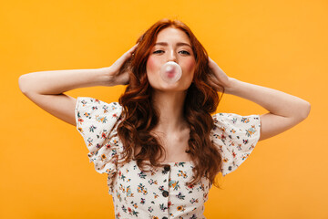 Close-up portrait of curly girl in white blouse with floral print. Woman making bubble of gum