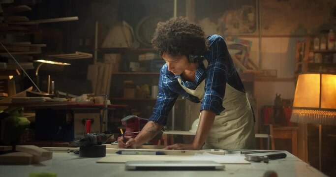 Handsome curly male carpenter in apron drawing design project at cozy workshop on desk, confident serious young man master using pencil at woodshop carpentry, craftsmanship, manufacturing concept