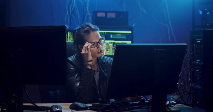 Caucasian good looking young woman in glasses, software developer or hacker working at the computer screen in the dark room late at night and being very tired, taking off glasses and giving her eyes a