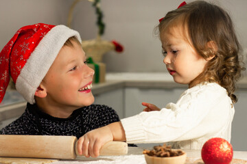 Little cute girl 2-4 with a red bow and boy 7-10 in a Christmas cap make gingerbread cookies in the New Year's kitchen.