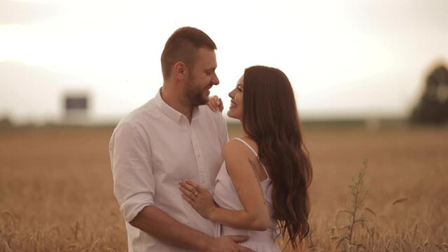 Chherful woman with long dark hair in white dress hugs with her boyfriend in white shirt on a field