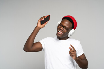 African American man with headphones wear red hat, enjoying listening to the artist's new album, dances, makes movements to music, holding mobile phone, isolated on studio gray background. 