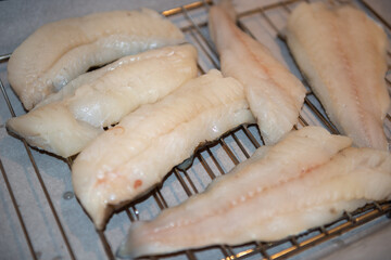 Multiple white wild codfish fillets on a metal rack drying. The raw fish is being prepared at a restaurant kitchen. The fresh uncooked cod cut into thick loin portions and whole pieces at a market.  
