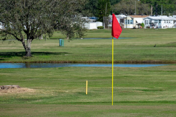 Obraz premium A red flag stands motionless attached to a yellow pole, at a golf hole, on a golf course. There are RV trailers in the background. There's a waterway, sand, and large trees near the putting hole