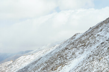 Snow covers rocky mountain slopes, which rise up into a sky filled with white clouds. Patches of...