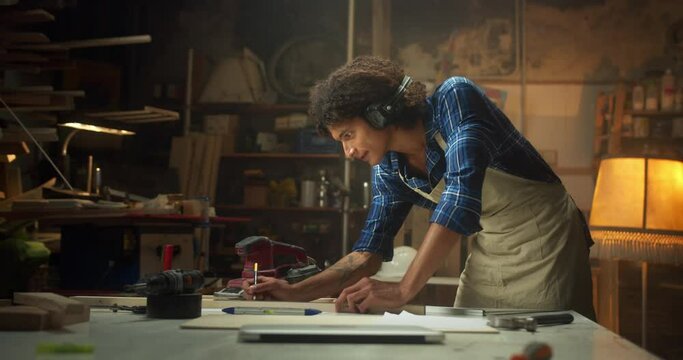 Side shot of pleasant carpenter drawing design carpentry project at cozy workshop indoors, attractive curly young man in apron and caged shirt leaning on work desk instruments engineer concept