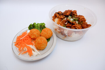 Japanese chicken rice, as known as Tori Don, is a chicken fillet cooked with japanese sauce placed on rice in a bowl. Tori don and three fish balls with vegetables beside it on a white background.