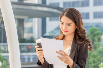 Fototapeta premium Happy business woman walking outside office with paper sheet and coffee cup in hand.
