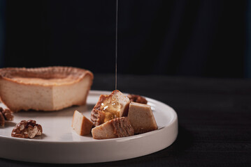 Small slices of ricotta cheese on white plate with nuts and honey at black background. A drop of honey falls into pieces of cheese.