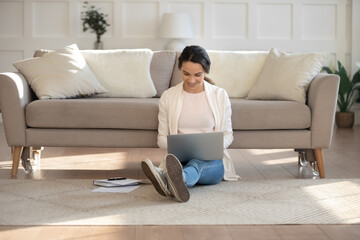 Working from home. Pleasant smiling young female student or freelancer sits on warm floor at living room holding laptop on knees studying, communicating with customers, taking part in video conference