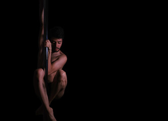 Dramatic dark portrait of a young man hanging from a pole dance bar with his eyes closed. Dark background studio shot.
