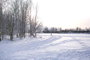 Winter landscape - white snow with traces of shoes and skis on the field. The ski track and road skirting the forest with bare trees, soft sunlight.