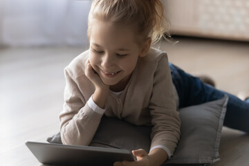 Close up smiling little girl using tablet, lying on warm wooden floor with underfloor heating at home, happy pretty child holding gadget, browsing apps, playing game, watching funny video