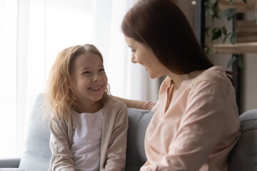 Overjoyed adorable little girl with loving young mother having fun at home, sitting on couch, caring mum hugging excited pretty daughter, chatting, talking, sharing secrets, spending leisure time