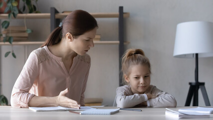 Strict mother scolding upset little daughter for bad marks, sitting at table at home, angry serious...