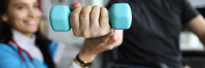 Close-up of physician female helping patient to develop joint. Man lifting blue dumbbell sitting in...
