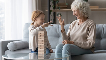Happy mature woman and little granddaughter playing funny board game, building towers from wooden blocks, sitting on cozy couch at home, elderly grandmother and laughing preschool girl having fun