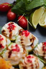 A set of sushi rolls lies on bamboo leaves with lime, herbs and red tomatoes, served on a black stone slate. Dark background