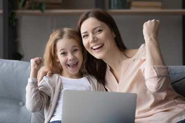 Head shot portrait overjoyed mother and little girl celebrating success, hugging, looking at camera, excited preschool girl and mum using laptop together, showing yes gesture, online lottery win