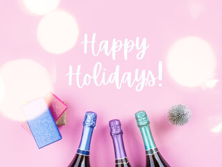 Champagne bottle, gifts and toy tree on pastel pink background. Festive flat lay with text Happy holidays.