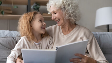 Close up smiling mature grandmother and little granddaughter reading book together, sitting on cozy...