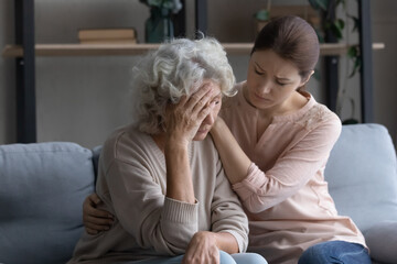 Caring grownup daughter comforting frustrated unhappy mature woman, hugging, sitting on couch at...