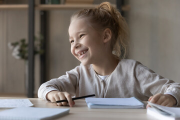 Naklejka premium Overjoyed motivated little girl studying, doing school homework, smiling pretty child schoolgirl looking to aside, dreaming and visualizing, distracted from assignments, homeschooling concept
