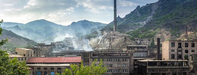 Panoramic view on old factory with half ruined buildings and exhaust fumes against green Caucasus...
